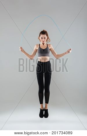 Full length portrait of a confident healthy fitness woman jumping with a skipping rope isolated over gray background
