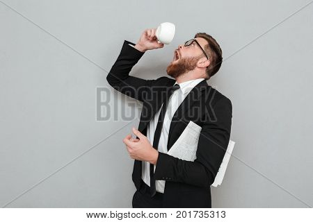 Funny young man in suit trying to drink last coffee drop from a cup isolated over gray background