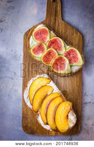 Toasted sandwiches with cream cheese topped with fresh ripe figs and peaches. Drizzled with honey. Wholegrain rye bran bread on wood cutting board grey concrete background
