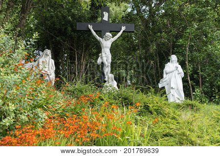 Clifden Ireland - August 4 2017: Calvary scenery with black cross and all white statues set against wood and in flower bed at Catholic Church.