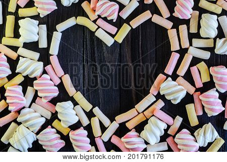 Laid out in the shape of heart multicolored marshmallows on black wooden table top view. Background of multi-colored and different in shape and size of marshmallows. Sweets background