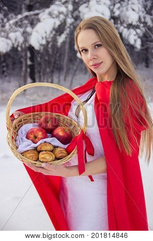 Girl in costume Little Red Riding Hood with dog like a wolf and a basket with apples and pies in the winter in the woods in the snow