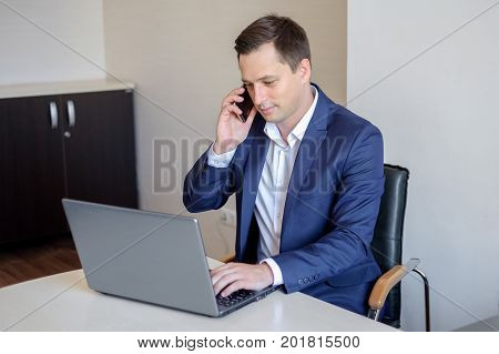 Smiling young businessman sitting behind his desk with laptop and talking on mobile phone in office.