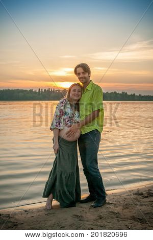 A pregnant beautiful woman with her husband on the river bank beach smiling and touching her mehandi ornament belly with love and care. Happy couple, family relax on sunset