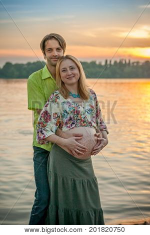 A pregnant beautiful woman with her husband on the river bank beach smiling and touching her mehandi ornament belly with love and care. Happy couple, family relax on sunset
