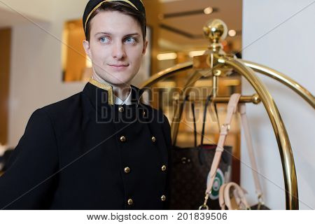 Smiling man in bellboy uniform standing with luggage cart and looking away.