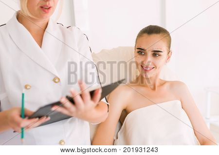 Doctor taking notes while patient is talking. Doctor or nurse talking to patient in cosmetology clinic. Woman patient smiling doctor.