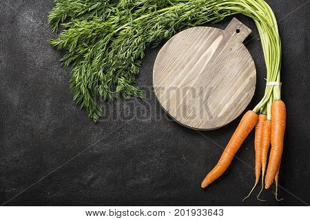 A bunch of fresh juicy carrots with a tops on a dark background with a vintage cutting board and a knife for cooking dinner. Top View