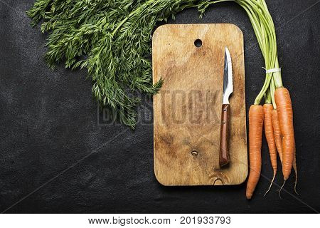 A bunch of fresh juicy carrots with a tops on a dark background with a vintage cutting board and a knife for cooking dinner. Top View