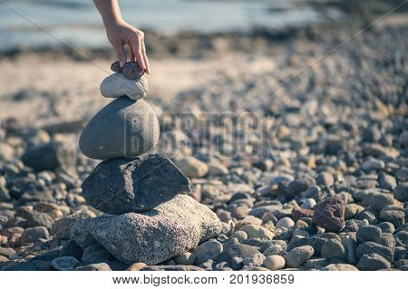 A female hand puts a small stone on top of a pyramid of stones. Stones on the beach.