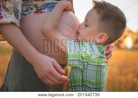 Cute boy kissing and listening his mothers pregnant mehandi pattern ornament belly on sunset nature landscape