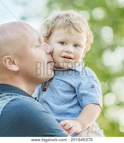 A happy family a young father kissing his adorable small son outdoor at summer park. Blurred background