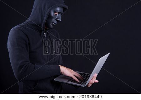 Anonymous male hacker using black mask to cover his face carrying laptop computer. Internet security and cyber attack concepts.