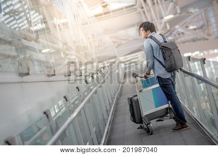 Young Asian man walking with airport trolley and his suitcase luggage in the international airport terminal arrival from travel abroad
