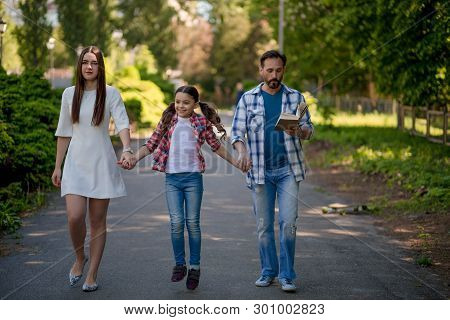 Family Walking Down The Road Outside In The City Park. Family Is Holding Each Other Hands. The Gaugh