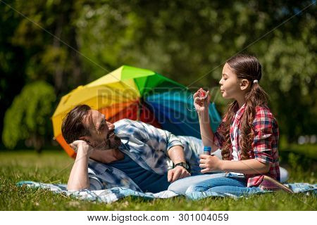 Father And Daughter On The Blue Blanket With Soap Bubbles In Park. Daughter Is Blowing Soap Bubbles 