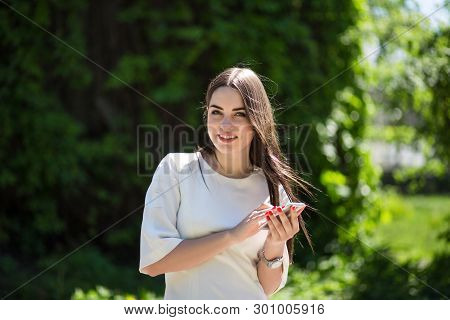 Smiling Young Brunette Woman Is Holding Her Phone In The Park. She Is Wearing A White Dress And A Si