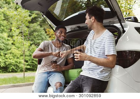 Young Multicultural Male Friends Sitting In Open Car Back, Drinking Coffee With Brofist Gesture