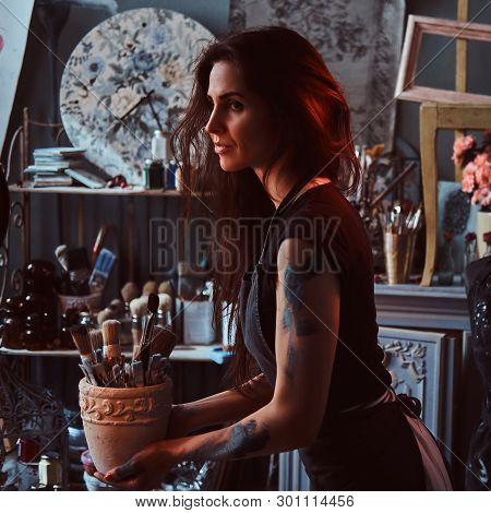Young Attractive Painter Is Holdind Jar With Brushes In Her Own Cozy Workshop.