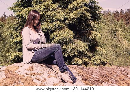 Young Pretty Girl Is Talking On The Phone While Sitting On A Large Stone Against The Background Of A