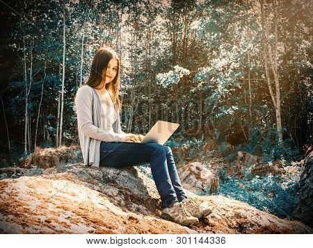 Young Beautiful Girl Is Working On A Laptop While Sitting On A Big Rock In The Forest. Freelance Con