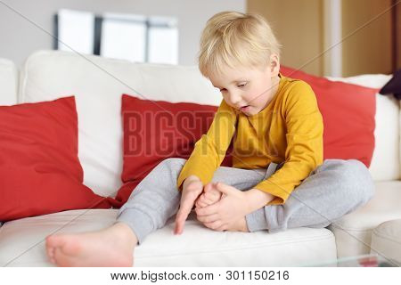 Little Boy Examines The Sore Leg Sitting On The Couch Indoors. Injury, Bruise, Splinter. Kids Trauma