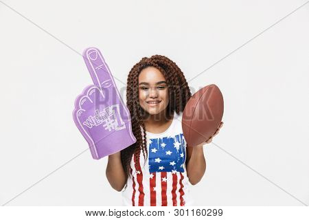 Portrait of joyous african american woman holding number one fan glove and rugby ball while standing isolated against white wall