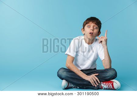 Image of smart brunette boy 10-12y with freckles wearing white casual t-shirt showing finger upward while sitting on floor isolated over blue background