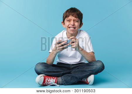 Image of smiling brunette boy 10-12y with freckles wearing white casual t-shirt holding and using smartphone while sitting on floor isolated over blue background