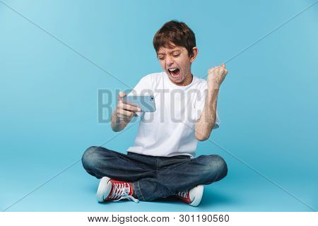 Image of ecstatic brunette boy 10-12y with freckles wearing white casual t-shirt holding and using smartphone while sitting on floor isolated over blue background