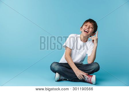 Image of thin brunette boy 10-12y with freckles wearing white casual t-shirt showing finger upward while sitting on floor isolated over blue background