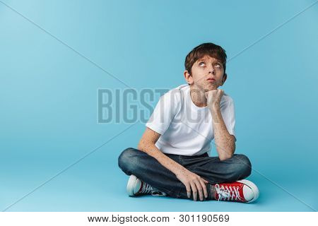 Image of european teenage boy 10-12y with freckles wearing white casual t-shirt looking upward while sitting on floor isolated over blue background
