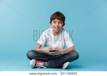 Image of european brunette boy 10-12y with freckles wearing white casual t-shirt holding and using smartphone while sitting on floor isolated over blue background