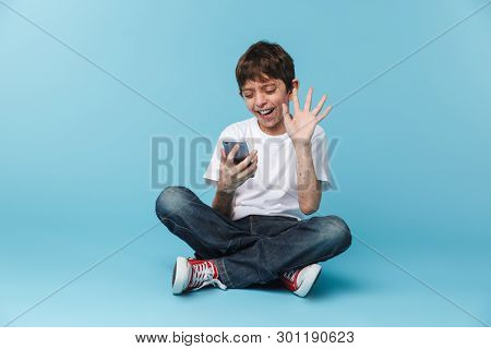 Image of european cheery boy 10-12y with freckles wearing white casual t-shirt holding and using smartphone while sitting on floor isolated over blue background