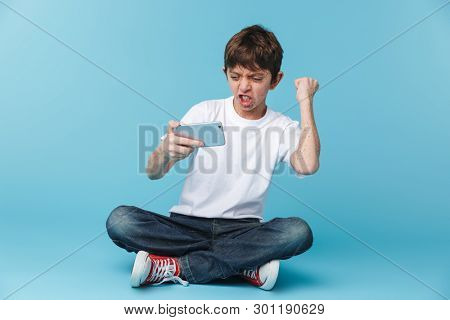 Image of happy brunette boy 10-12y with freckles wearing white casual t-shirt holding and using smartphone while sitting on floor isolated over blue background