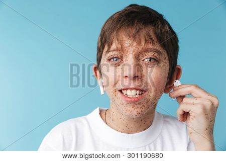 Image of funny caucasian boy 10-12y with freckles wearing white casual t-shirt and earpods looking at camera isolated over blue background