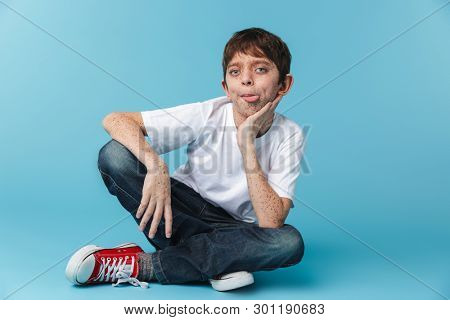 Image of funny brunette boy 10-12y with freckles wearing white casual t-shirt looking at camera while sitting on floor isolated over blue background