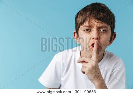 Image of young boy 10-12y with freckles wearing white casual t-shirt holding index finger on lips isolated over blue background