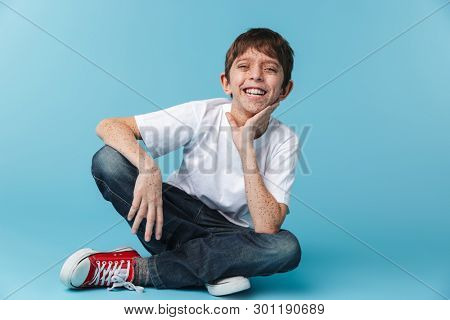 Image of joyful brunette boy 10-12y with freckles wearing white casual t-shirt smiling at camera while sitting on floor isolated over blue background