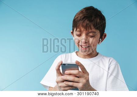 Image of joyful caucasian boy 10-12y with freckles wearing white casual t-shirt smiling and holding smartphone isolated over blue background