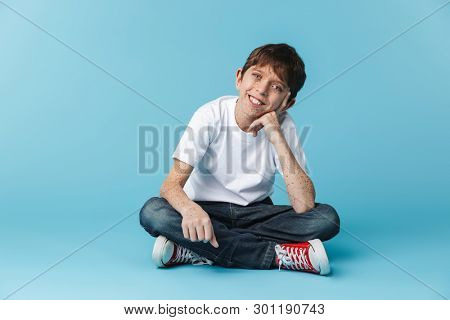 Image of european brunette boy 10-12y with freckles wearing white casual t-shirt smiling at camera while sitting on floor isolated over blue background