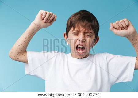 Image of emotional caucasian boy 10-12y with freckles wearing white casual t-shirt and earpods screaming at camera isolated over blue background