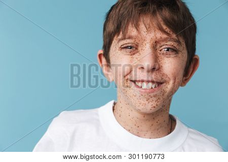 Portrait closeup of beautiful youngster boy 10-12y with freckles wearing white casual t-shirt smiling at camera isolated over blue background