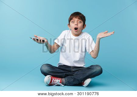 Image of european excited boy 10-12y with freckles wearing white casual t-shirt holding and using smartphone while sitting on floor isolated over blue background