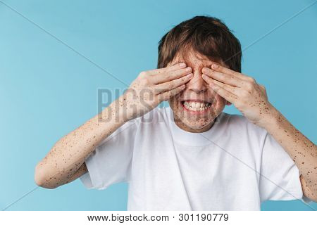 Image closeup of cheery caucasian boy 10-12y with freckles wearing white casual t-shirt having fun isolated over blue background