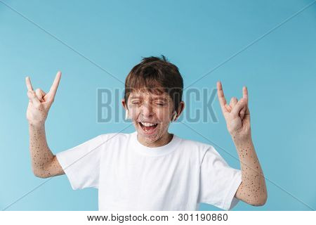Image of happy caucasian boy 10-12y with freckles wearing white casual t-shirt and earpods showing rock sign camera isolated over blue background
