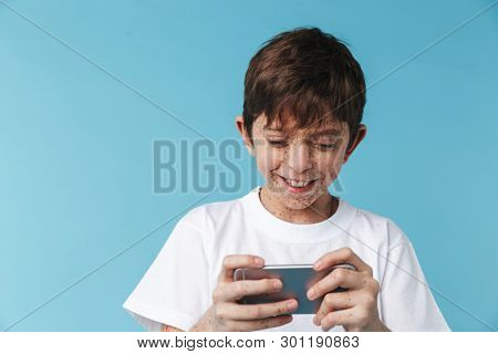 Image of pleased caucasian boy 10-12y with freckles wearing white casual t-shirt smiling and holding smartphone isolated over blue background