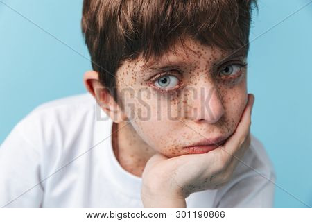 Portrait closeup of young beautiful boy 10-12y with freckles wearing white casual t-shirt looking at camera isolated over blue background