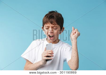 Image of young caucasian boy 10-12y with freckles wearing white casual t-shirt and earpods singing at camera isolated over blue background