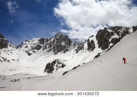 Snow Covered Mountain Range With Avalanche Traces, Sunlit Cloudy Sky And Hiker In Red With Dog On Sn